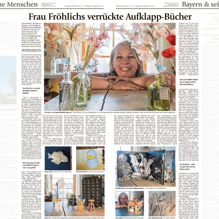 Lächelnde Frau (Julia Schreiber) an einem Tisch mit Papierblumen und geöffneten Büchern.